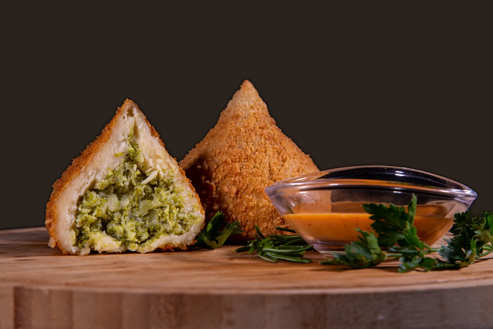 Coxinha de brócolis aberta ao lado de uma inteira, com molho, numa tábua de madeira
