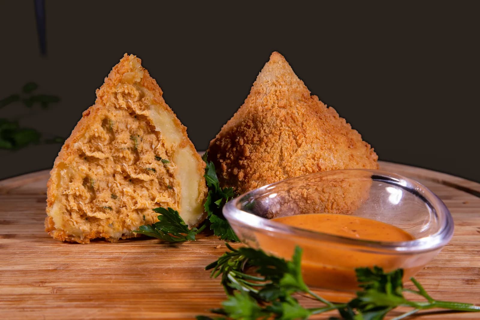 Coxinha de frango com requeijão aberta ao lado de uma inteira, com molho, numa tábua de madeira