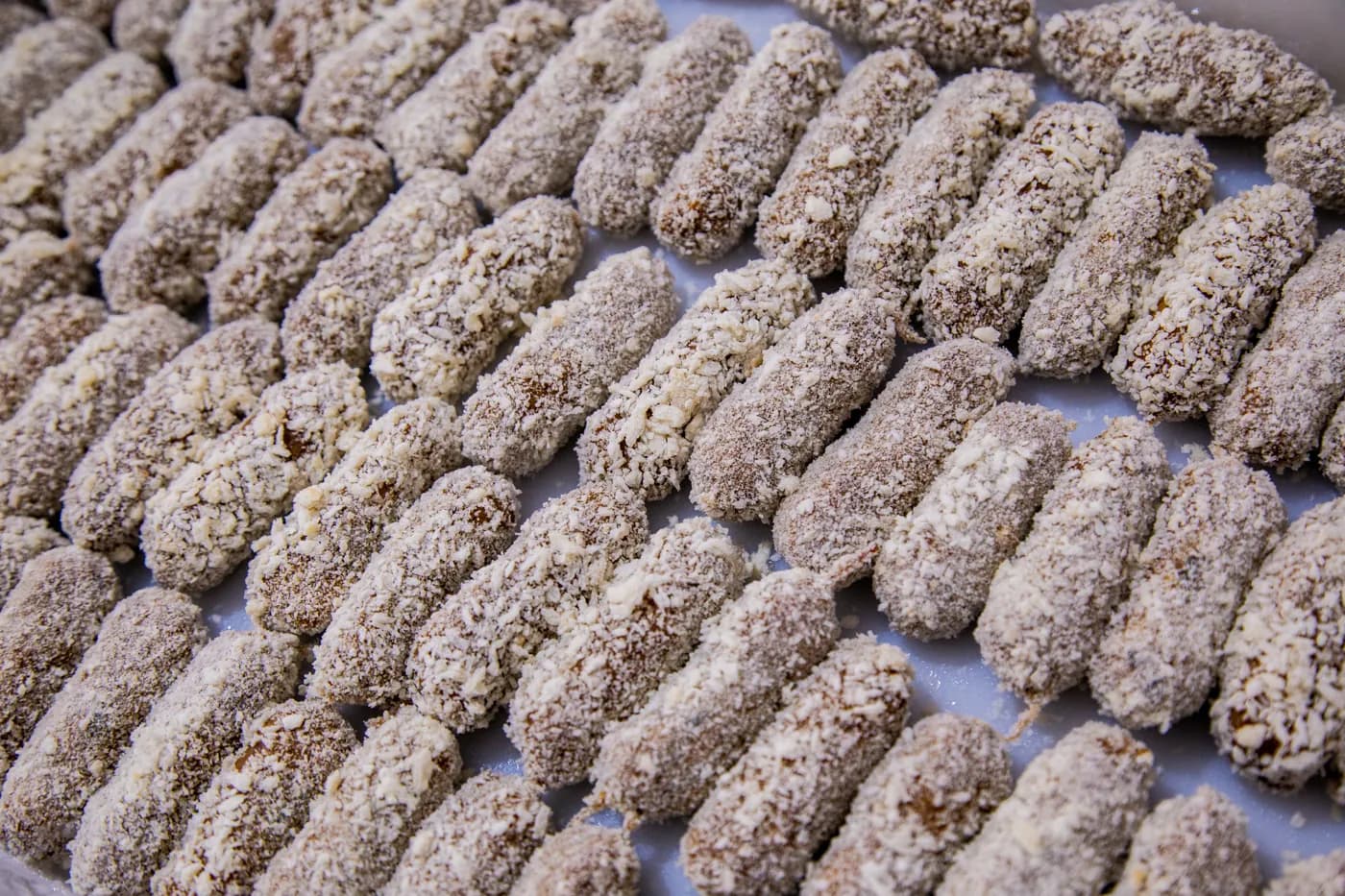 Salgados empanados crus, compridos, enfileirados numa bandeja